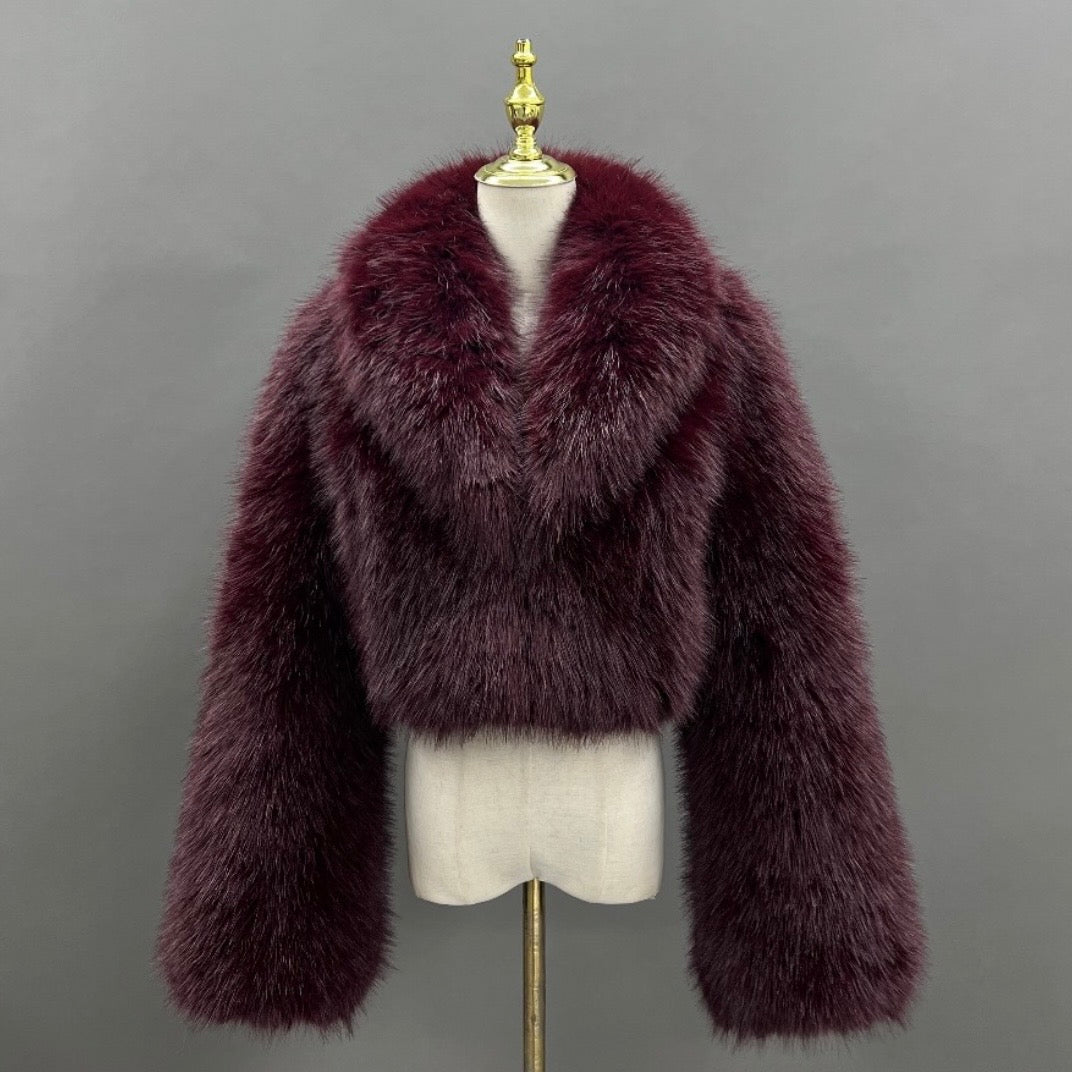 A plush, cropped fur jacket in a deep burgundy or plum shade, displayed on a cream-colored mannequin torso with a gold finial against a solid gray background.